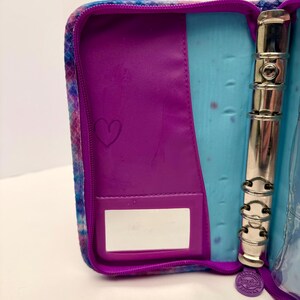 May include: Open, zippered binder with a purple interior and a tie-dye exterior in shades of blue, purple, and pink. The interior features a purple pocket with a heart drawn on it, a clear pocket, and a silver, three-ring binder mechanism.