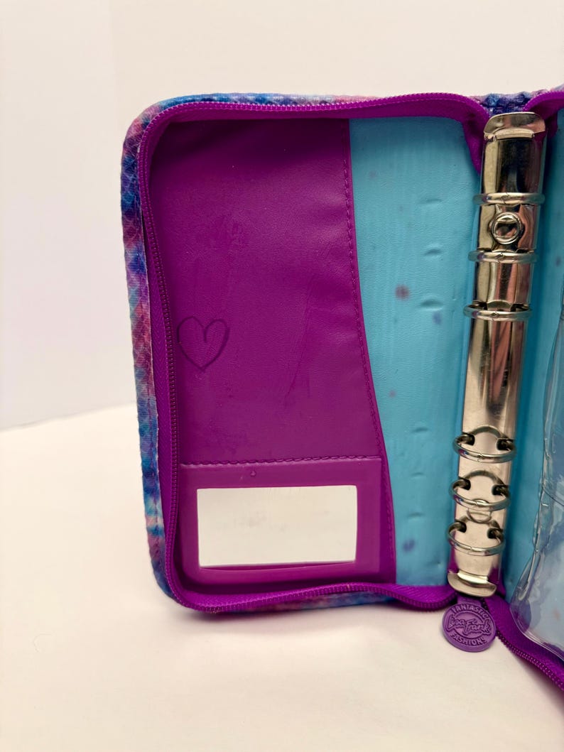 May include: Open, zippered binder with a purple interior and a tie-dye exterior in shades of blue, purple, and pink. The interior features a purple pocket with a heart drawn on it, a clear pocket, and a silver, three-ring binder mechanism.