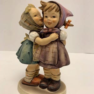 Könnte beinhalten: Keramikfigur, die zwei junge Mädchen beim Umarmen zeigt. Eines küsst das andere auf die Wange. Sie tragen Kleider, Schürzen und Kopftücher. Die Figur steht auf einem runden, cremefarbenen Sockel.