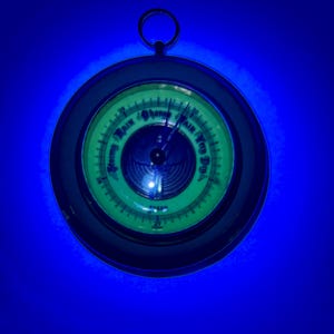 May include: A vintage, round barometer with a dark frame and a green face. The face displays text indicating weather conditions like "Stormy," "Rain," and "Fair." A needle points to the current atmospheric pressure. The barometer has a small ring at the top.