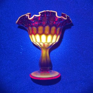 May include: A glass compote dish featuring a deep red and amber gradient color scheme. The bowl has a ruffled, crimped rim and a vertical ribbed pattern along the sides. It stands on a pedestal base with a circular foot, glowing under UV light.