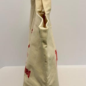 K&ouml;nnte beinhalten: Cremefarbene Canvas-Tragetasche mit roter Schrift. Die Tasche hat zwei Griffe und einen rechteckigen Boden. Der Text auf der Tasche ist teilweise sichtbar. Die Tasche wird von einer Person gehalten.