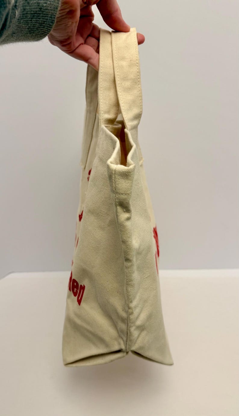 K&ouml;nnte beinhalten: Cremefarbene Canvas-Tragetasche mit roter Schrift. Die Tasche hat zwei Griffe und einen rechteckigen Boden. Der Text auf der Tasche ist teilweise sichtbar. Die Tasche wird von einer Person gehalten.