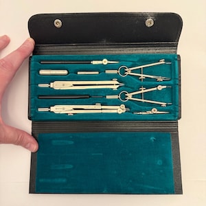 May include: A black leatherette folding case containing a set of silver-toned metal drafting compasses and technical drawing tools. The interior is lined with teal velvet, featuring molded slots to secure each individual instrument in place.