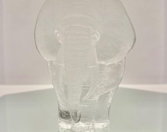 May include: A clear glass elephant sculpture. The elephant is depicted in a frontal pose, with detailed features including large ears, a trunk, and tusks. The sculpture is displayed on a reflective surface.