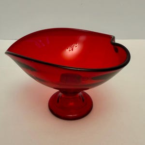 Puede incluir: Un cuenco de vidrio rojo vibrante con un borde negro, que descansa sobre una base de pedestal. El cuenco tiene una forma asimétrica única. Esta pieza decorativa es adecuada para servir o exhibir.