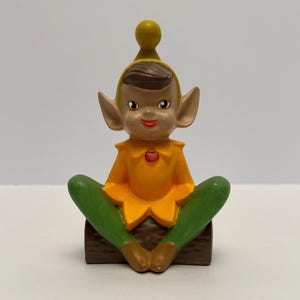 Peut inclure: Figurine d'elfe en céramique avec un chapeau et une chemise jaunes, un pantalon vert et des chaussures marron. L'elfe a de grandes oreilles, un visage souriant et un cœur rouge sur la chemise. L'elfe est assis sur une bûche brune.