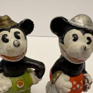 May include: Two vintage Mickey Mouse figurines. One Mickey wears a gold-colored hat and green pants, the other wears a silver-colored hat and red pants. Both have black ears and arms, white faces, and red smiles.