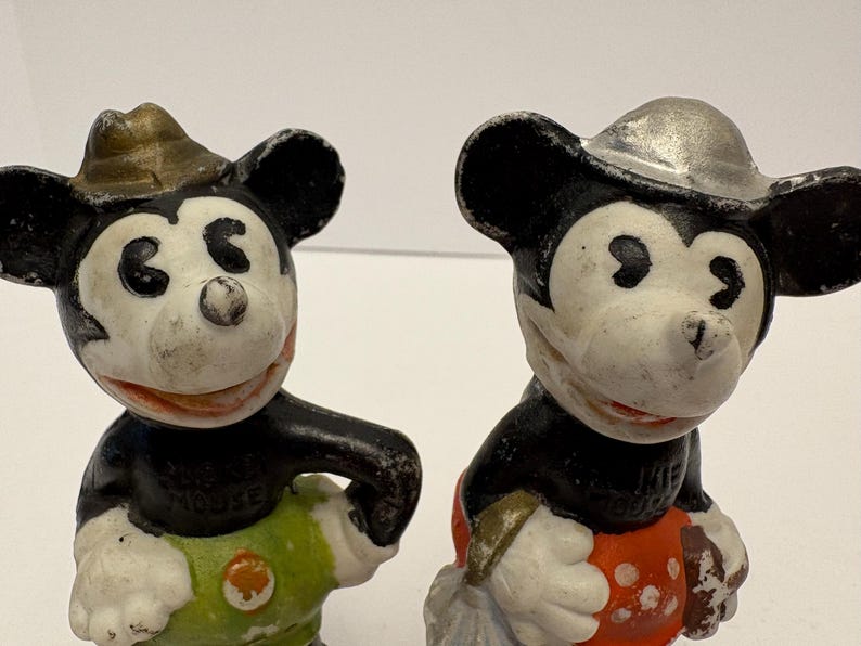 May include: Two vintage Mickey Mouse figurines. One Mickey wears a gold-colored hat and green pants, the other wears a silver-colored hat and red pants. Both have black ears and arms, white faces, and red smiles.