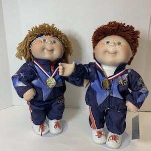 Puede incluir: Dos muñecas Cabbage Patch Kids vestidas con chándales azules y blancos con detalles en rojo. Cada muñeca lleva una medalla y zapatillas blancas con cordones rojos. Una muñeca tiene el pelo rubio y una diadema, la otra tiene el pelo rojo.