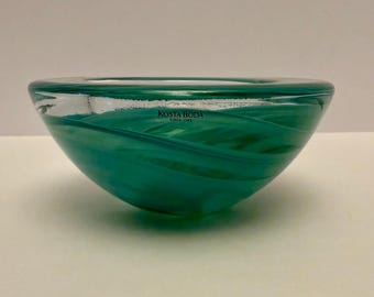 May include: A teal-colored glass bowl with a swirling design. The bowl has a rounded shape and a clear rim. The text "KOSTA BODA SINCE 1742" is visible on the side. This decorative bowl is suitable for home decor.
