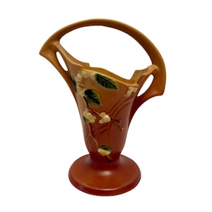 Pode incluir: Um vaso de cerâmica castanho com uma pega, com um design floral com folhas verdes e flores brancas. O vaso tem uma abertura larga e uma base redonda, adequado para exibir flores ou como peça decorativa.