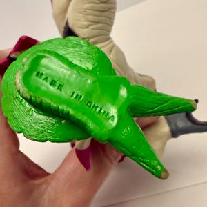 May include: A green plastic figurine with the text "MADE IN CHINA" stamped on the bottom. The figurine has a textured surface and is held by a hand. The figurine is part of a larger toy.
