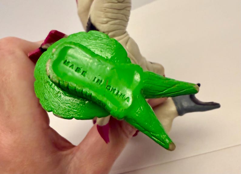 May include: A green plastic figurine with the text "MADE IN CHINA" stamped on the bottom. The figurine has a textured surface and is held by a hand. The figurine is part of a larger toy.