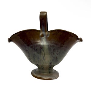 Op de afbeelding: Een keramische mand met een handvat, met een uitlopende, geschulpte rand. De mand heeft een voetstuk en is geglazuurd in tinten bruin, groen en wit. De handgreep is effen bruin van kleur. De algemene vorm doet denken aan een bloem.