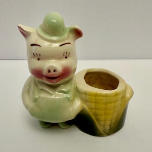 Könnte beinhalten: Ein Vintage-Keramik-Pflanzgefäß in Form eines lächelnden Schweins mit grünem Hut und Latzhose. Das Schwein hat rosige Wangen und hält einen gelben Korb. Der Pflanztopf ist ein charmantes Dekorationsobjekt.
