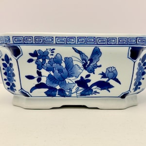May include: A rectangular ceramic planter featuring a white base with blue floral and butterfly motifs. The rim is decorated with a geometric Greek key pattern, and the sides display detailed blue painted flowers and a butterfly design.