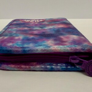 May include: A rectangular wallet with a tie-dye pattern in shades of purple, blue, and white. The wallet has a purple zipper and a round zipper pull with the text "FANTASTIC FASHIONS".