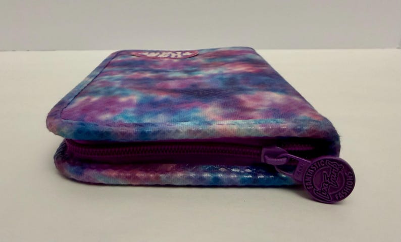 May include: A rectangular wallet with a tie-dye pattern in shades of purple, blue, and white. The wallet has a purple zipper and a round zipper pull with the text "FANTASTIC FASHIONS".
