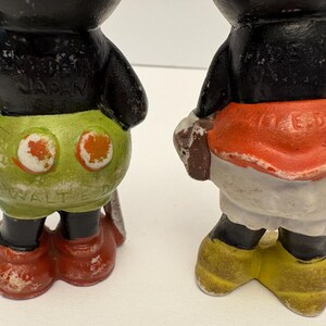 May include: Two vintage Mickey Mouse figurines. One has green shorts with orange circles and red shoes. The other has a red skirt, white shorts, and yellow shoes. Both have black bodies and are weathered.