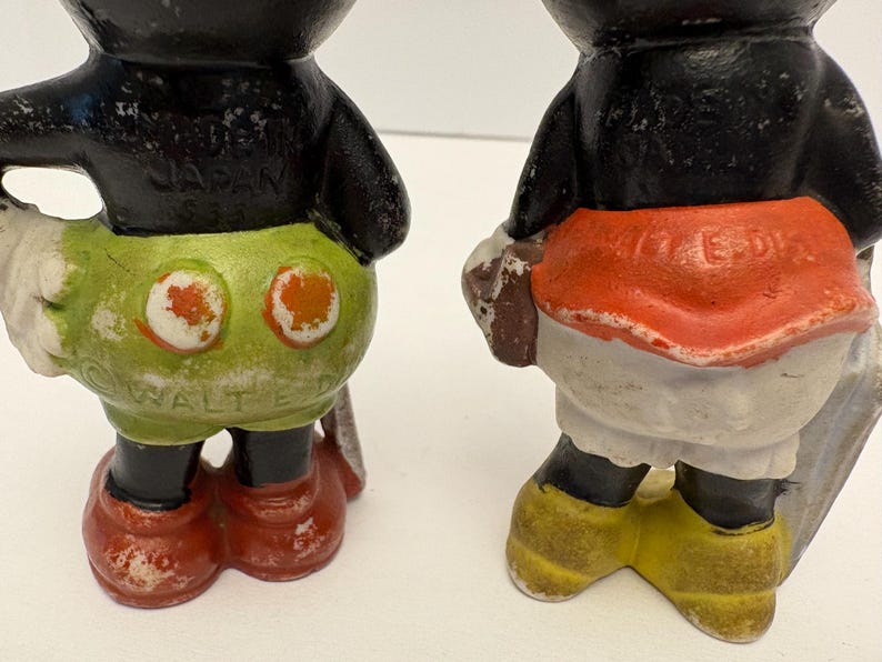 May include: Two vintage Mickey Mouse figurines. One has green shorts with orange circles and red shoes. The other has a red skirt, white shorts, and yellow shoes. Both have black bodies and are weathered.