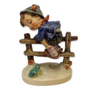May include: A ceramic figurine of a young person climbing over a wooden fence. The figure wears a blue jacket, green hat, and patterned socks. A small frog sits at the base of the fence, and red flowers are scattered around.