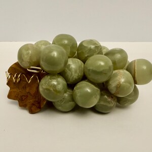 May include: A decorative cluster of green marble grapes. The grapes are round and smooth, with varying shades of green. They rest on a carved wooden base with a gold-colored metal accent.