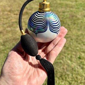 Può includere: Un flacone di profumo vintage con una base rotonda bianca e un design a righe blu e bianche. Presenta una parte superiore color oro e un atomizzatore nero con nappa. Il flacone è tenuto in una mano.