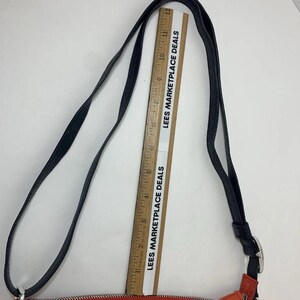 May include: Black and red crossbody bag with a silver buckle and decorative emblem. The bag has a long black strap and orange accents. The text "LEES MARKETPLACE DEALS" is printed on a ruler.