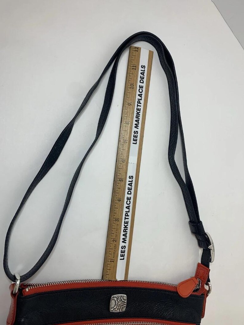 May include: Black and red crossbody bag with a silver buckle and decorative emblem. The bag has a long black strap and orange accents. The text "LEES MARKETPLACE DEALS" is printed on a ruler.