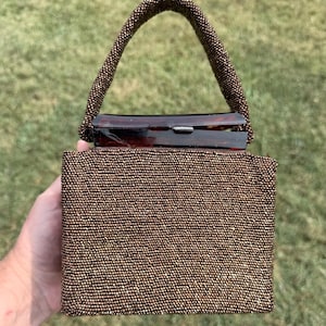 Op de afbeelding: Een vintage handtas met kralen in bruine tinten. De rechthoekige tas heeft een met kralen bezette handgreep en een schildpadsluiting. De tas wordt tegen een groene achtergrond gehouden.