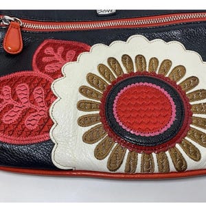 May include: Black leather crossbody bag with a red zipper and pull. The bag features a large, embroidered flower design in white, brown, red, and black. Additional red embroidered leaf accents are on the side. The bag has a red trim.