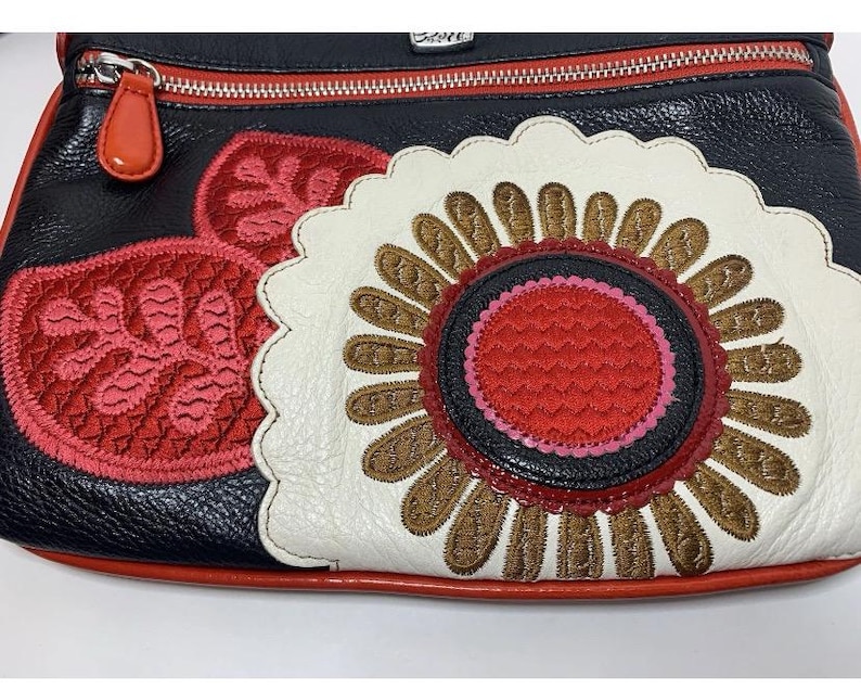 May include: Black leather crossbody bag with a red zipper and pull. The bag features a large, embroidered flower design in white, brown, red, and black. Additional red embroidered leaf accents are on the side. The bag has a red trim.
