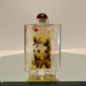 Peut inclure: Une bouteille en verre transparent avec un bouchon marron, ornée d'une scène peinte de deux grues, d'un soleil rouge et d'oiseaux en vol. La bouteille est encastrée dans un support rectangulaire transparent.