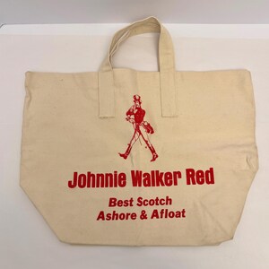 K&ouml;nnte beinhalten: Cremefarbene Canvas-Tragetasche mit rotem Text und einer Grafik eines gehenden Mannes. Der Text lautet "Johnnie Walker Red", "Best Scotch" und "Ashore & Afloat". Die Tasche hat zwei Griffe.