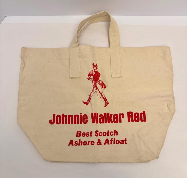 K&ouml;nnte beinhalten: Cremefarbene Canvas-Tragetasche mit rotem Text und einer Grafik eines gehenden Mannes. Der Text lautet "Johnnie Walker Red", "Best Scotch" und "Ashore & Afloat". Die Tasche hat zwei Griffe.