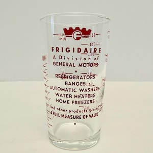 以下が含まれることがあります： 赤色の文字と目盛りの付いた透明なガラス製メジャーカップ。「FRIGIDAIRE A Division of General Motors」と書かれ、冷蔵庫やレンジなどの家電製品が記載されています。カップにはカップと液量オンスの測定線があります。