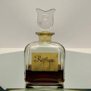 May include: A vintage perfume bottle with a clear glass body and a decorative stopper. The bottle contains a dark amber liquid and features a label with the text "Replique" and "Raphael Paris". The stopper is shaped like a stylized tulip.