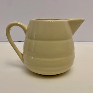 May include: A cream-coloured ceramic jug with a rounded body and a slightly flared spout. The jug has a simple, elegant design with horizontal lines around the body.
