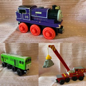 Könnte beinhalten: Eine Sammlung von drei Spielzeugeisenbahnen, bestehend aus einer violetten Lokomotive mit der Aufschrift Charlie und der Nummer 14 sowie roten Rädern, einer grünen Lokomotive mit der Aufschrift D5702 und einem roten Kranwagen mit gelb-rotem Ausleger.