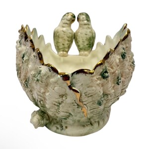 Puede incluir: Un cuenco decorativo de cerámica con diseño de concha, con dos pequeñas figuras de pájaros. El cuenco es principalmente blanco con detalles verdes y dorados. Los pájaros son de color verde claro con detalles dorados.
