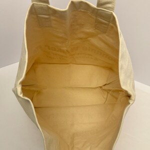 K&ouml;nnte beinhalten: Eine beigefarbene Canvas-Tragetasche mit rechteckiger Form und zwei Henkeln. Der Innenraum ist hellbeige. Die Tasche wird offen gehalten und zeigt das Innere. Die Tasche ist aus robustem Material gefertigt.