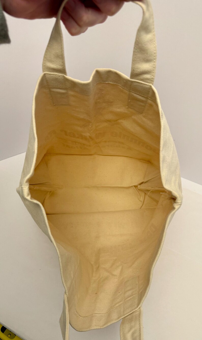 K&ouml;nnte beinhalten: Eine beigefarbene Canvas-Tragetasche mit rechteckiger Form und zwei Henkeln. Der Innenraum ist hellbeige. Die Tasche wird offen gehalten und zeigt das Innere. Die Tasche ist aus robustem Material gefertigt.