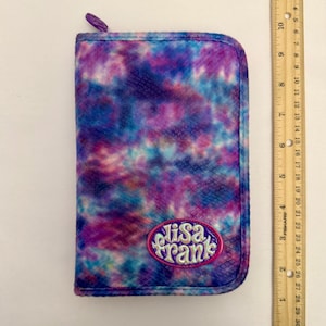 May include: A rectangular Lisa Frank pencil case with a tie-dye pattern in shades of purple, blue, and pink. The case has a zipper closure and a Lisa Frank logo patch. A ruler is next to the case.