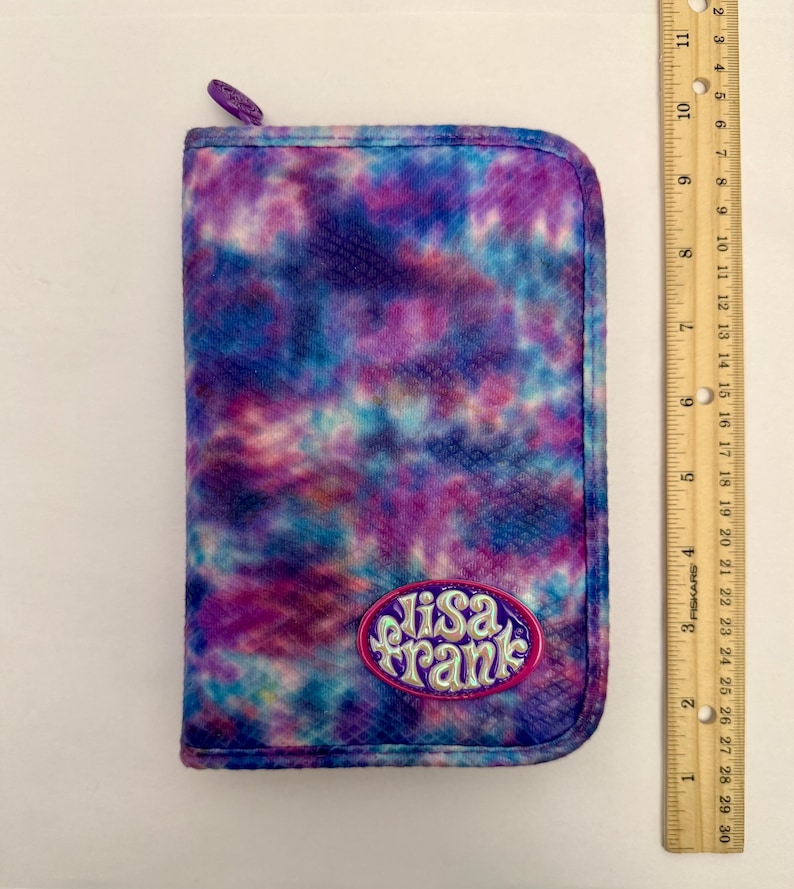 May include: A rectangular Lisa Frank pencil case with a tie-dye pattern in shades of purple, blue, and pink. The case has a zipper closure and a Lisa Frank logo patch. A ruler is next to the case.