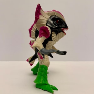 May include: A colorful action figure of a creature with a black head, red eyes, and a wide open mouth. The figure has a cream-colored body with pink and black accents, green legs, and gray arms. The figure is posed in a dynamic stance.