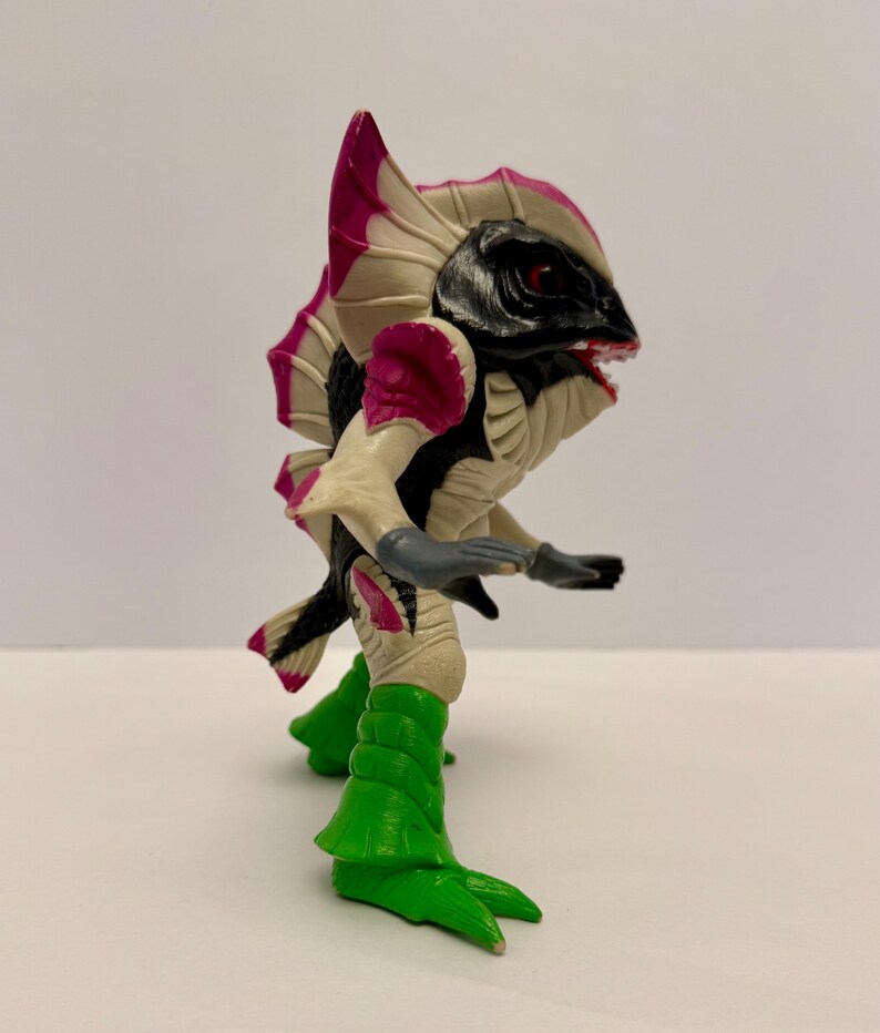 May include: A colorful action figure of a creature with a black head, red eyes, and a wide open mouth. The figure has a cream-colored body with pink and black accents, green legs, and gray arms. The figure is posed in a dynamic stance.