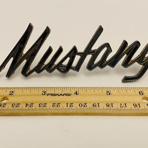 May include: A vintage "Mustang" emblem in a cursive font, approximately 6 inches long, with a dark metallic finish. A wooden ruler with inch markings is placed below the emblem.
