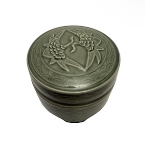 May include: A round, celadon ceramic container with a lid. The lid features a raised floral design with two stylized flowers and a bird. The container has a textured surface and a darker band around the middle. The design is elegant and detailed.
