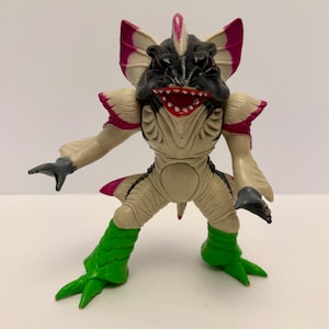 May include: A detailed figurine of a creature with a black head, a wide red mouth, and large, fan-like appendages with pink accents. The figure has tan skin, green legs, and gray hands, showcasing a unique design.